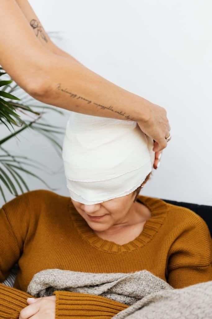 A woman with a bandaged head is tended to by a caring person indoors.