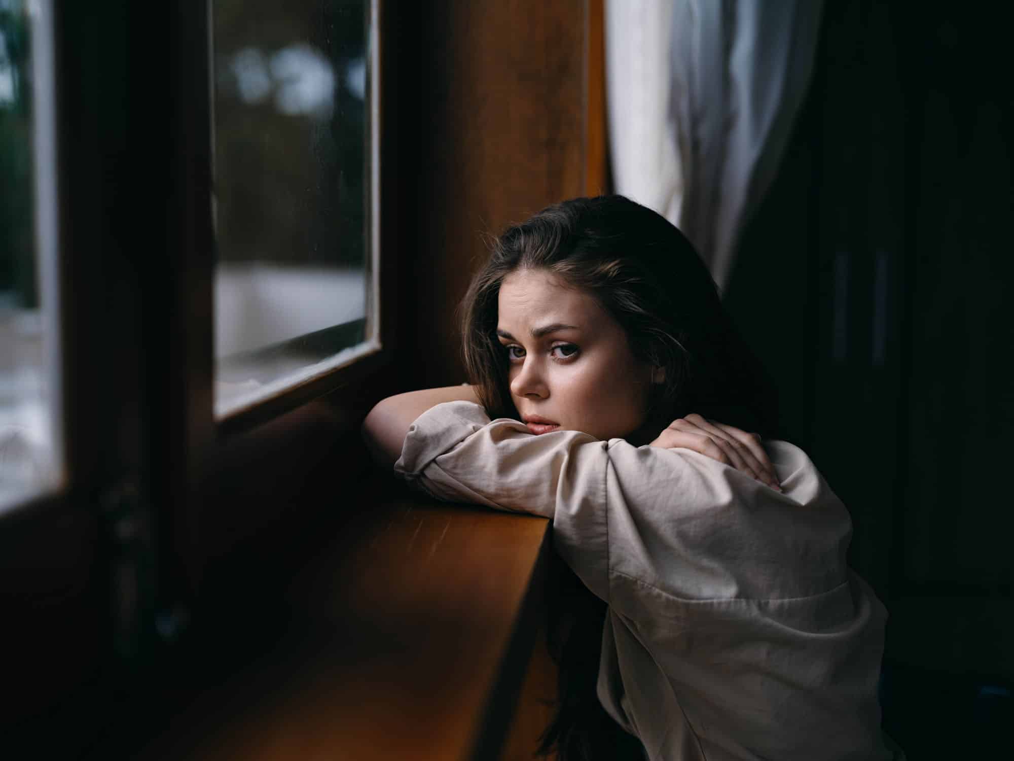 Portrait of young woman at home by window with wooden frame, autumn mood, cozy lifestyle, sleepy weather.